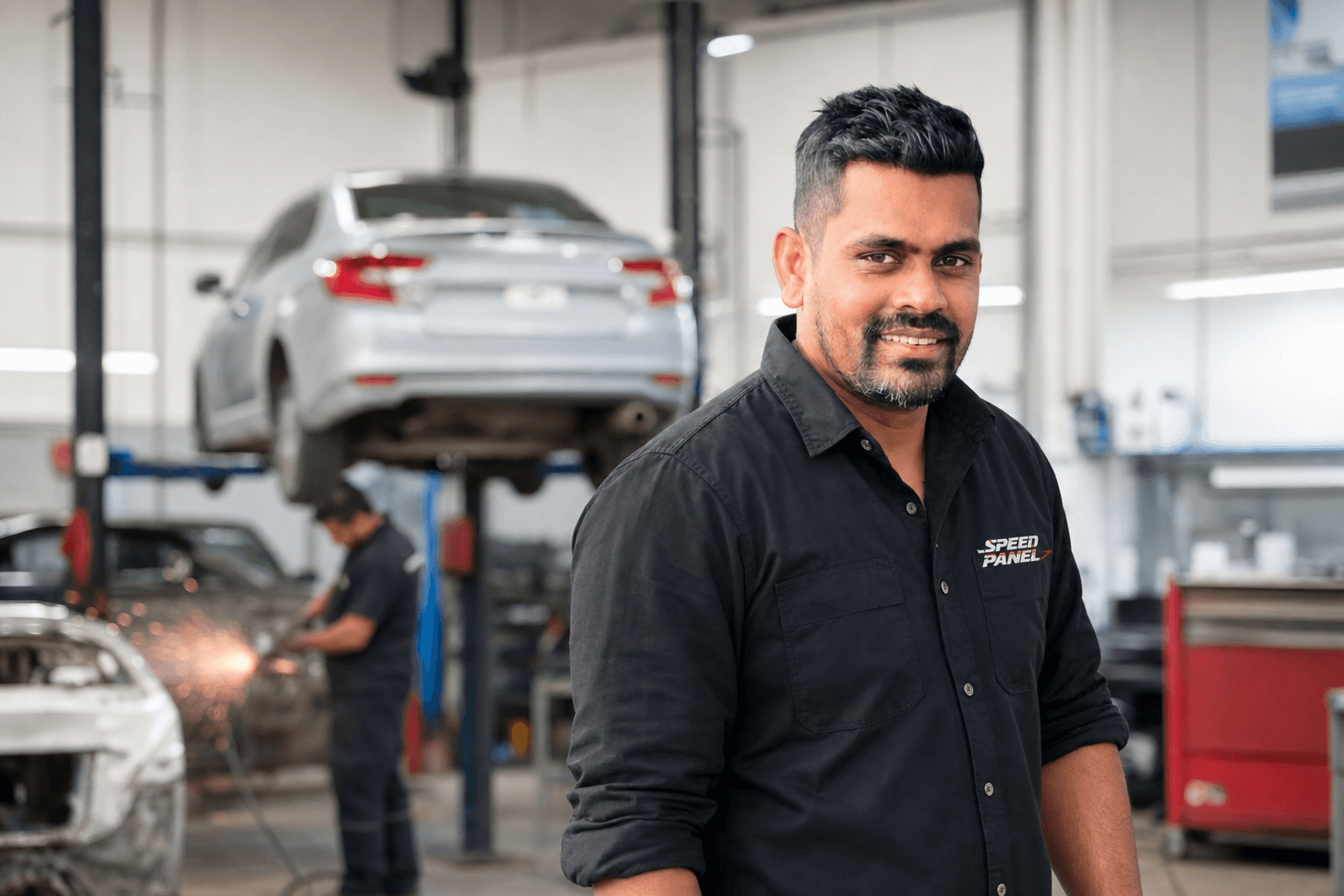 Dinesh, owner of Speed Panel & Paint, in workshop uniform with active panel repairs in the background